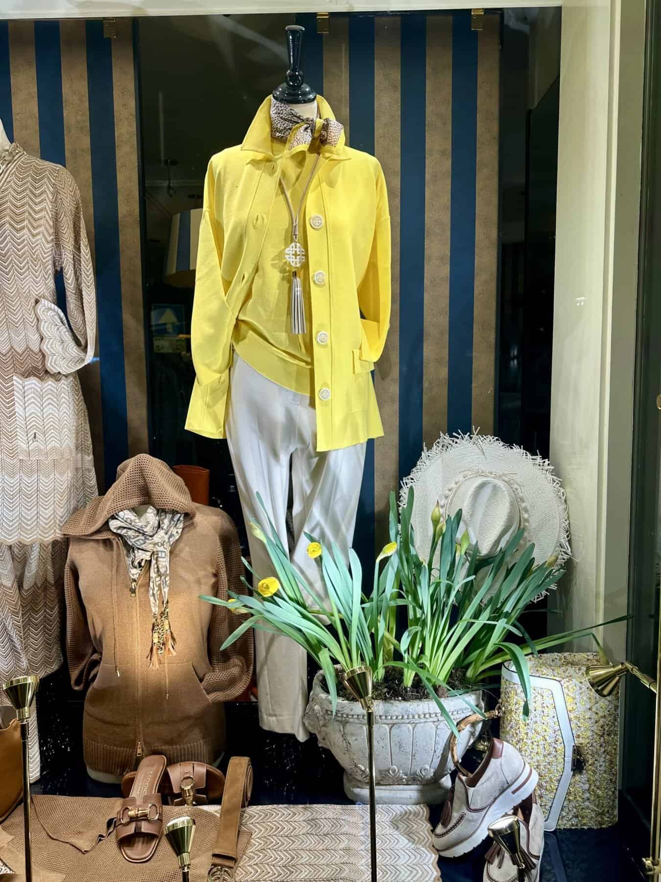 Modici winkelraam met mannequin in gele jas en witte broek, accessoires en decoratieve planten.